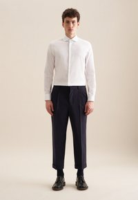 Camicia bianca con bottoni abbinata a pantaloni blu con pieghe e scarpe di pelle nere, creando un look smart-casual con una vestibilità su misura.