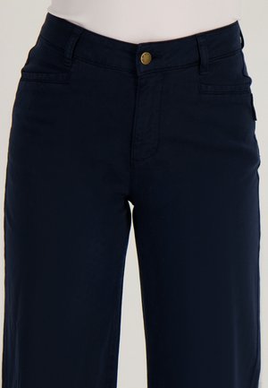 Vrouw die donkerblauwe rechtgesneden broek draagt met knopen aan de voorkant en zakken, gecombineerd met een wit shirt dat is ingestopt.