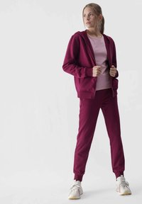 Bordeauxfarbener Reißverschluss-Hoodie, passende Jogginghose und hellrosa T-Shirt. Das Material scheint weich zu sein mit gerippten Bündchen und einer lässigen Passform. Weiße Sneakers.