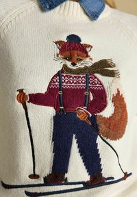 Krēmkrāsas adīta džemperis ar izšūtu lapsu, kas valkā bordo džemperi, zilus bikses un rakstainu šalli, tur rokās slēpošanas nūjas.