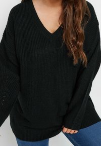 Kvinna som bär en svart kraftig stickad V-ringad tröja med långa ärmar och blå jeans, med vågigt brunt hår över ena axeln.