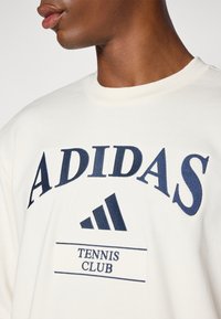Balta sporta krekls ar jūras zilas krāsas "ADIDAS" logo un treknu uzrakstu "TENNIS CLUB" virs trīs svītru Adidas emblemas.