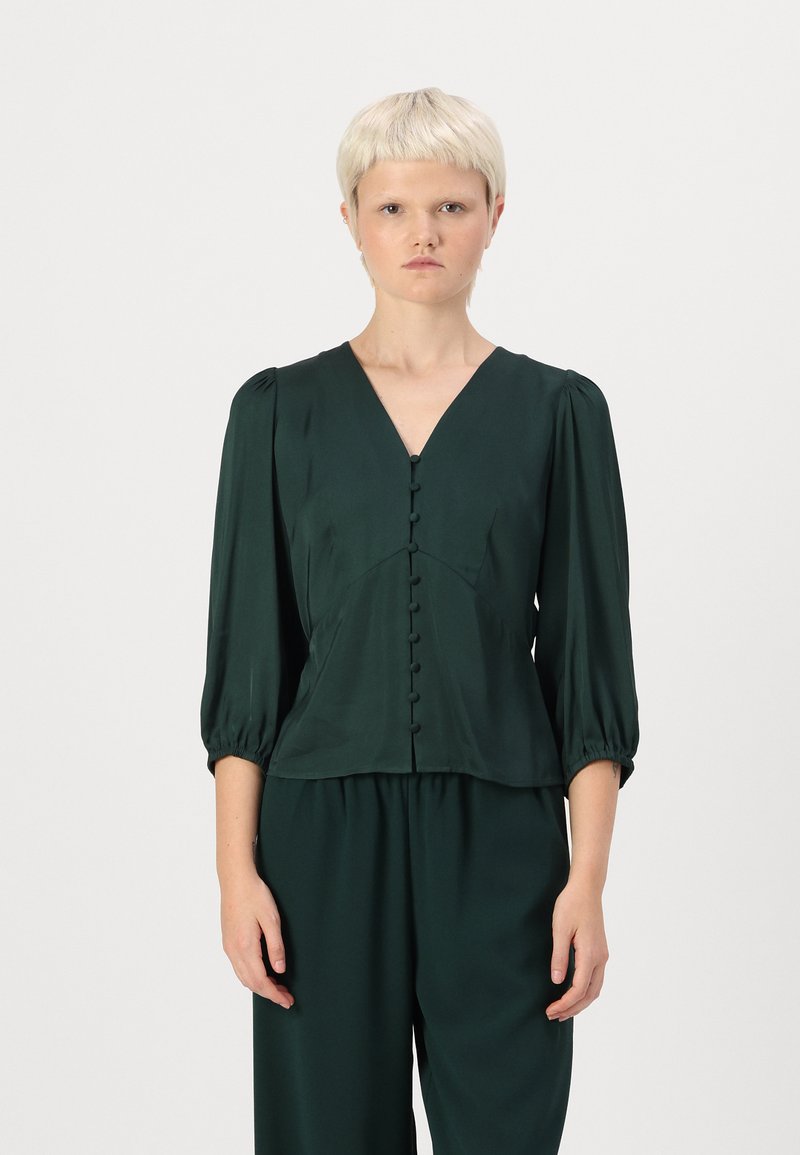 Mørkegrønn bluse laget av et glatt stoff, med V-hals, puffede ermer og en rad med knapper nedover fronten.