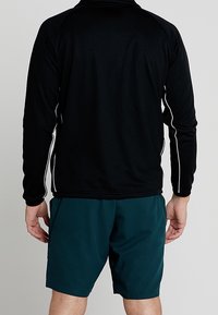 Persona che indossa una maglietta nera a maniche lunghe con strisce bianche sui lati e pantaloni corti verde scuro, in piedi con la schiena rivolta verso l'osservatore.