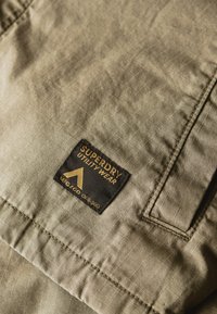 Khaki stof met een rastertextuur, voorzien van een rechthoekig zwart etiket met de tekst "SUPERDRY UTILITY WEAR" en een logo in het geel.
