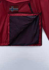 Rode windjack met een grote gaaszak. Bevat een kleine bruine patch met een blauw-wit vlagontwerp. Glad aanvoelend.