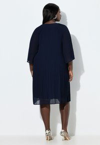 Granatowa plisowana sukienka z rękawami do łokcia, o linii A i przezroczystej fakturze; zestawiona z wysokimi obcasami w wzór węża.