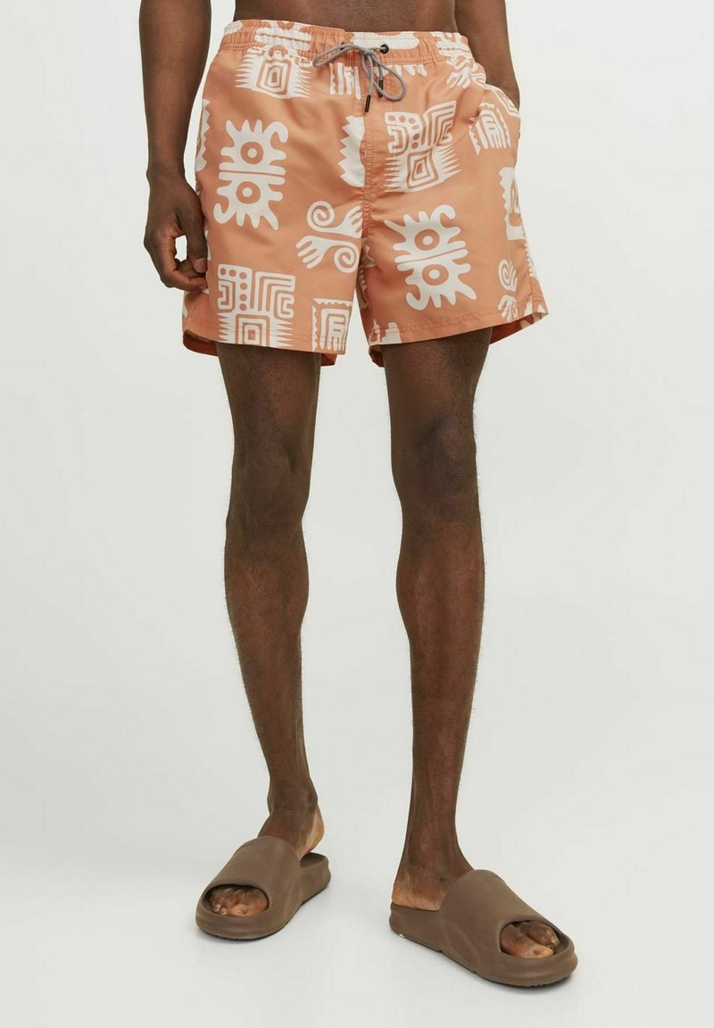 Zwemshorts in licht perzikkleur met witte geometrische patronen, voorzien van een elastische tailleband en een trekkoord. Draag ze met bruine instapsandalen.