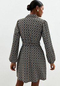 Mujer con cabello oscuro en un moño, vistiendo un vestido de manga larga con cinturón y un patrón de diamantes en negro, azul y beige, de espaldas contra un fondo liso.