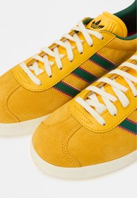 Κίτρινα σουέντ αθλητικά παπούτσια με πράσινες και κόκκινες ρίγες, λευκά κορδόνια και υφασμένο καουτσούκ σόλα. Επένδυση στη γλώσσα.