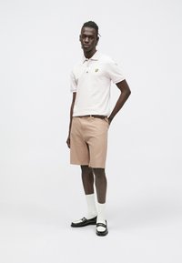 Lichtgekleurde poloshirt met een geel logo, gecombineerd met beige gestreepte korte broek. Draagt witte sokken en zwarte loafers.