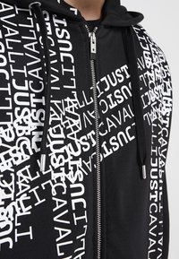 Hoodie zippé noir avec texte blanc "JUST CAVALLI" imprimé en diagonale et se chevauchant sur le devant et les manches, avec des cordons noirs.