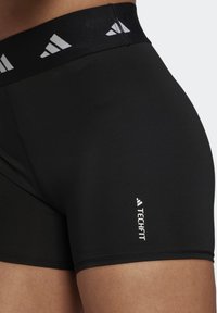 Mustat urheilushortsit, joissa on leveä elastinen vyötärönauha, jossa on harmaa geometrinen kuosi; "TECHFIT"-logo valkoisena sivussa. Sileä pinta.