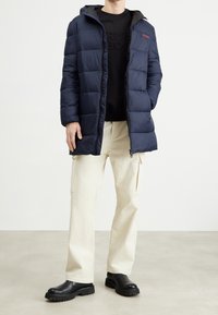 Chaqueta acolchada navy con capucha, textura acolchada, bolsillos laterales y logo rojo en el pecho, combinada con pantalones cargo beige y botas negras.