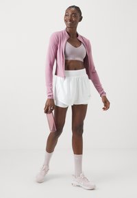Chaqueta atlética de color rosa claro sobre un sujetador deportivo gris claro, combinado con pantalones cortos blancos. Sosteniendo una botella de agua rosa, con zapatillas y calcetines a juego.