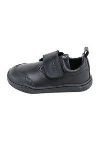 Zapato sintético negro con puntera redondeada, correa de velcro, capucha texturizada en la punta y suela plana de goma. Incluye una pestaña en el talón para facilitar el calzado.