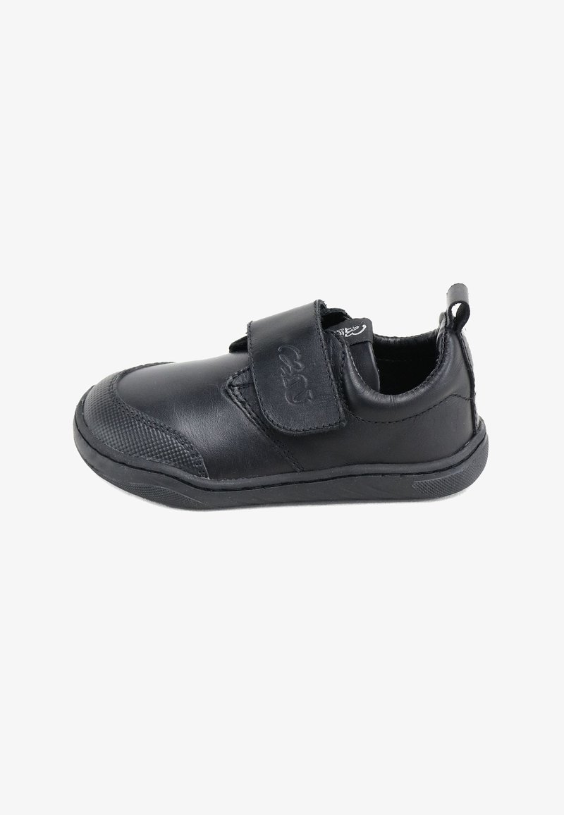 Zapato sintético negro con puntera redondeada, correa de velcro, capucha texturizada en la punta y suela plana de goma. Incluye una pestaña en el talón para facilitar el calzado.