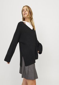 Černý oversized svetr s výstřihem do V, s kleslými rameny, žebrovanými manžetami a bočními rozparky, nošený přes bílou košilovou část.