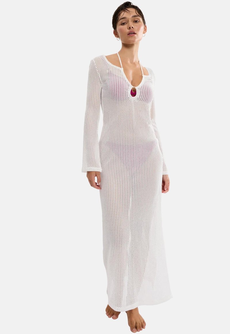 Γυναίκα που φοράει ένα μακρύ, λευκό, ανοιχτό πλεκτό φόρεμα πάνω από ένα ροζ μαγιό, όρθια ξυπόλητη μπροστά σε ένα απλό φόντο.