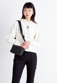 Felpa crema con cappuccio e cordino, caratterizzata da un piccolo logo. Borsa a tracolla nera con spallacci. Il modello indossa pantaloni neri.