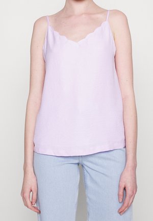 Person iført en lys lilla ærmeløs top med bølgede kanter og lyseblå jeans, vist fra skuldrene til hofterne.