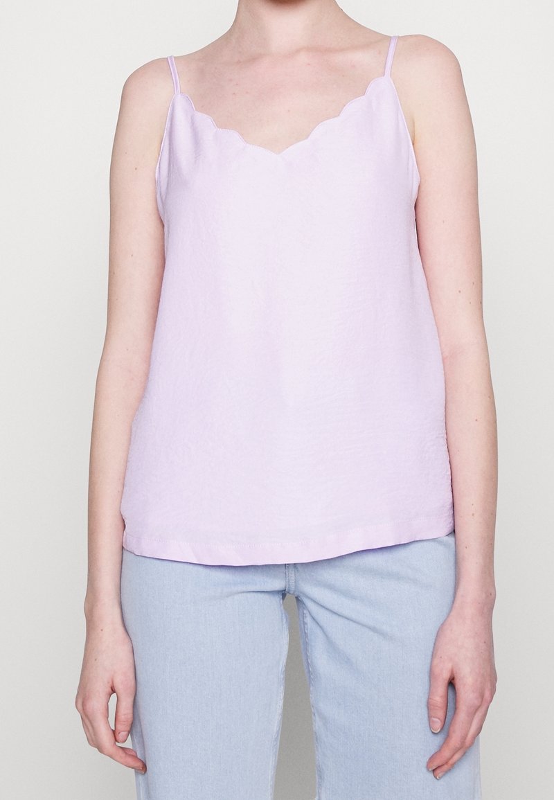 Osoba ubrana w jasnofioletowy top bez rękawów z falowanym dekoltem oraz jasnoniebieskie dżinsy, pokazywana od ramion do bioder.