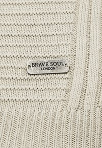 Tkanina z pletené béžové příze s texturovaným vzorem a stříbrným kovovým štítkem s vyraženým nápisem "BRAVE SOUL" a "LONDON."