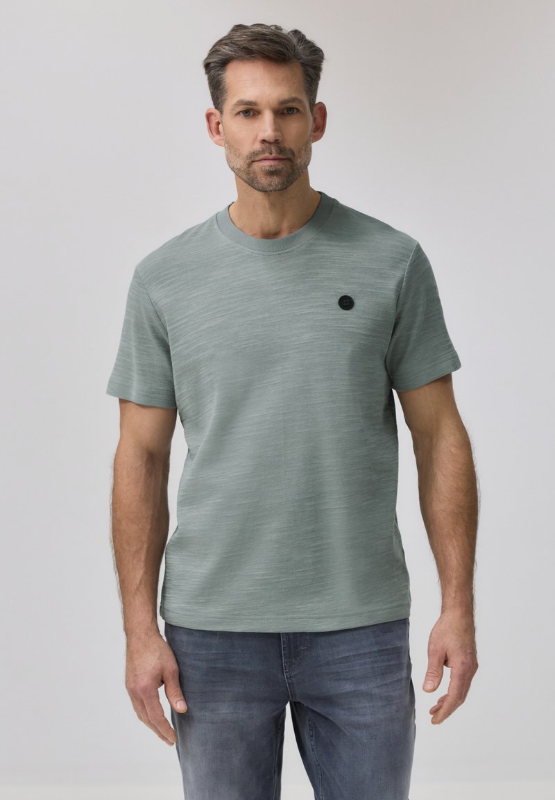 Homme aux cheveux courts et foncés et à la barbe taillée portant un t-shirt vert clair texturé et un jean bleu délavé sur un fond uni.