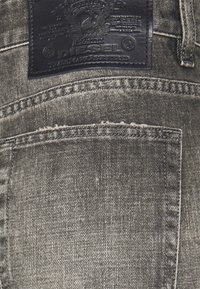 Denimkangas tummanharmaassa pesussa, jossa on ommeltu takatasku ja teksturoitu musta nahkapatch, jossa on kohokuvioitu "Diesel"-logo.