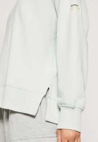 Světle zelená mikina se žebrovanými manžetami a lemem. Má boční rozparky blízko lemu a malou značkovou etiketu na horním rukávu. Měkká textura.