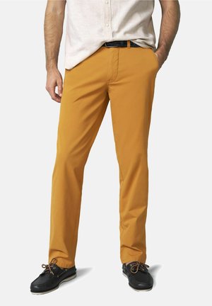 Mann trägt senfgelbe Freizeithose, schwarzen Gürtel, beige kurzärmliges Hemd und schwarze Bootsschuhe, steht mit den Händen in den Taschen.