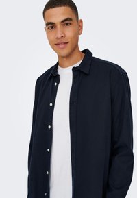 Temno modra srajca z gumbi in ovratnikom, izdelana iz teksturirane tkanine z belimi gumbi, nošena čez belo majico.