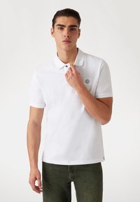 Weißes Polohemd aus einer Baumwollmischung, mit klassischem Kragen, kurzen Ärmeln und einem Logo-Patch auf der Brust. Vorgestellt mit dunkelgrünen Hosen.