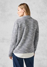 Texturerad grå och marinblå herringbone-mönstrad jacka, med krage och långa ärmar, bärs över en vit skjorta och matchas med mörka jeans.