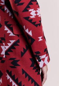 Manteau rouge à motifs avec des designs géométriques noirs et blancs, en laine, avec une poche unique et trois boutons noirs sur le poignet.