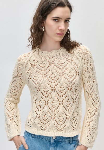 Νεαρή γυναίκα με σγουρά μαλλιά, φορεί ένα πουλόβερ μπέζ με ανοιχτό δίχτυ και μπλε τζιν, στέκεται με το χέρι στην τσέπη μπροστά σε απλό φόντο.