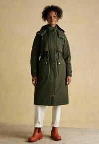 Długa oliwkowozielona kurtka puchowa z kapturem, z wcięciem w talii, dwoma przednimi kieszeniami i zapięciem na zatrzaski. Połączona z białymi spodniami i brązowymi botkami za kostkę.