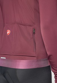 Aktív sportdzseki sötét burgundi színű anyagból, sima felülettel. Zipzáras zsebbel és kontrasztos, puha derékpánttal rendelkezik. Logófelvarrás található rajta.