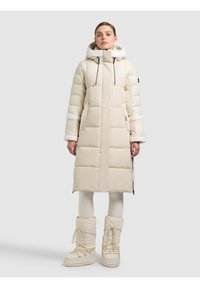 Kvinna klädd i lång beige vadderad kappa med huva och matchande vinterkängor, stående mot en ljusgrå bakgrund.