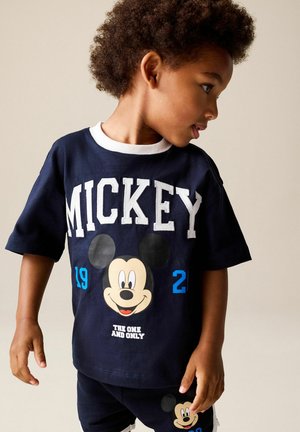 Barn iført et navyblåt sæt med Mickey Mouse T-shirt og shorts, kigger til siden mod en ensfarvet beige baggrund.