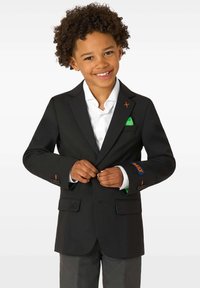 Lastehooldas lokkis juustega laps naeratab, samal ajal kui on nööpide nööpimisega musta bleiseri valge särgi, hallide pükste ja rohelise taskuäriga.