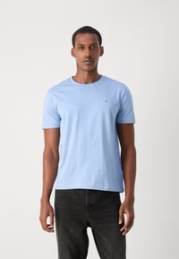 Light blue cotton t-shirt with short sleeves, crew neck, and a small logo. Paired with dark jeans in a fitted style.