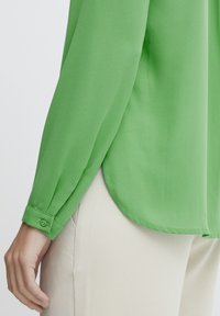 Green, long-sleeved blouse with a smooth texture, a curved hem, and a buttoned cuff. Worn over light-colored pants, showcasing the sleeve detail.