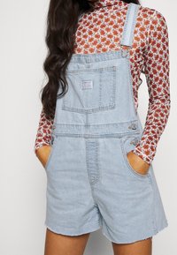 Tergal de mezclilla azul claro con tirantes ajustables, bolsillo frontal y botones laterales, usado sobre una blusa de flores de manga larga con cuello alto.