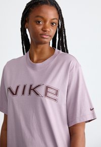 Camiseta de algodón de color púrpura claro con un gran logo de "NIKE" en púrpura oscuro en la parte frontal, manga corta y escote redondo.