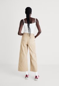 Béžové kalhoty s širokými nohavicemi a hladkým povrchem, se dvěma zadními kapsami a jemným prošitím. V kombinaci s bílým topem a teniskami s červenými detaily.