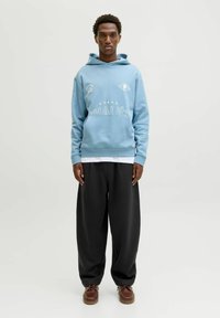 Lysblå hoodie med frontlomme og hvide tekstdetaljer, parret med løse sorte bukser og brune sko. Minimalistisk stil, glat tekstur.