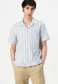 Camisa de botones de manga corta en azul claro con rayas verticales negras, con cuello muesca y logotipo bordado en el pecho.