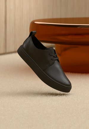 Czarny skórzany casualowy sneaker z sznurowadłami, unoszący się nad beżowym dywanem, z bursztynowym, zaokrąglonym szklanym stolikiem w tle.