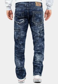 Jeans in denim di colore blu scuro con effetti di scolorimento e whiskering. Presentano più tasche con cuciture bianche e un'etichetta del marchio sulla vita.